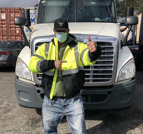 Man wearing a neon yellow jacket and a face mask, giving us a thumbs up and a rock n roll sign with his hands, standing in front of a white semi-truck, in a container yard