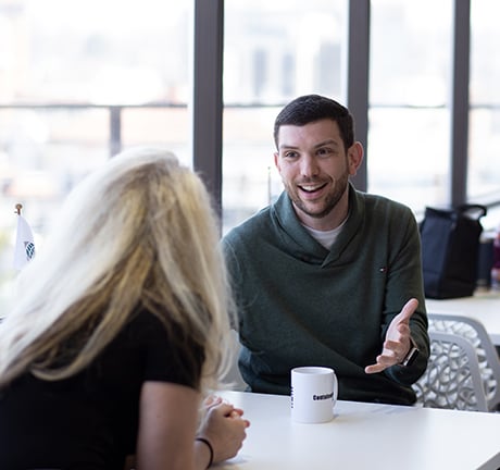 A white man is facing the camera and talking to a woman with blonde hair. He is pointing a finger at himself and smiling. Behind him is a woman looking at her smart phone while eating lunch.