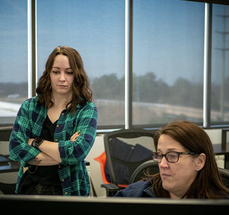 two women, one wearing a green flannel standing behind the other woman who is sitting behind a computer monitor, in an office