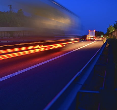 Image of a blurred shipping truck on a highway driving at night. Dark blue sky in the background, bright red headlights blurred from fast movement.