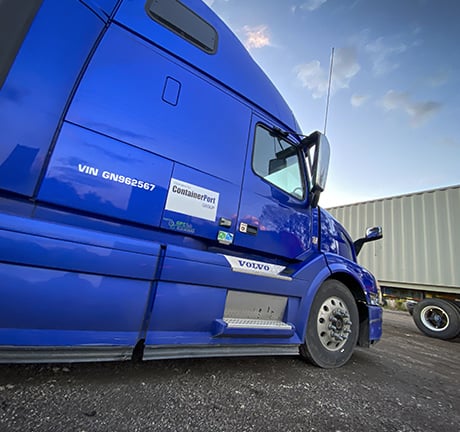 Blue shipping truck showing the passenger side, parked in front of a white shipping container